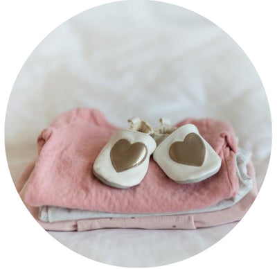 Light pink baby clothes folded on the bed. White baby shoes with gold hearts on on top.